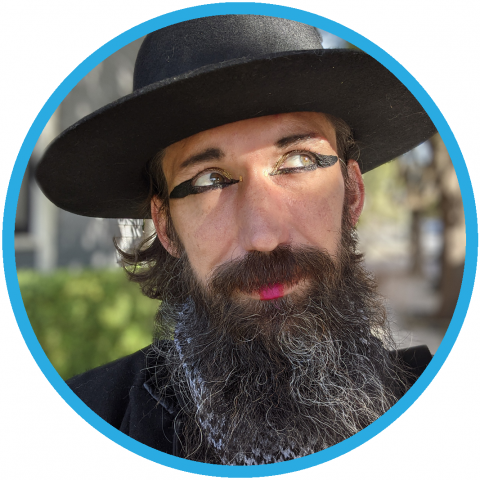 Jason Wyman is caucasian, male presenting queer person. They are wearing a black wide brimmed hat and has a long dark beard.