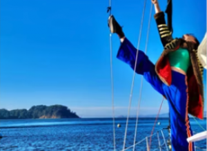 Circus performer standing on edge of sailing boat, one leg raised vertical up over their head, ocean is in the background