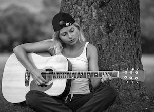 Female playing acoustic guitar. She is wearing a white tank top shirt and black baseball cap while sitting in front of a tree.
